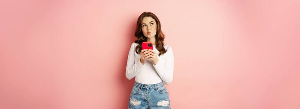 Online shopping. Glamour girl using smartphone to order in app internet, holding mobile phone, thinking, and smiling, looking up thoughtful, standing over pink background. - Photo, image