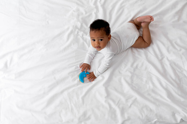 Top View tiro de bebê preto bonito brincando com brinquedo de Rattle enquanto deitado na cama em casa, adorável menino afro-americano pequeno ou menina relaxante em lençóis brancos no quarto e olhando para a câmera - Foto, Imagem