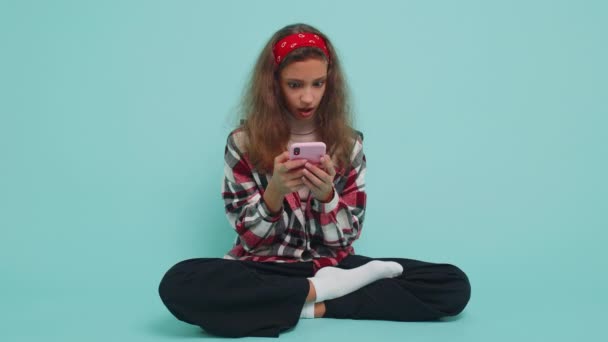Young teenager child girl kid use mobile smartphone typing browsing say Wow yes found out great big win good news celebrate doing winner gesture. Happy preteen children sitting on blue wall background - Footage, Video