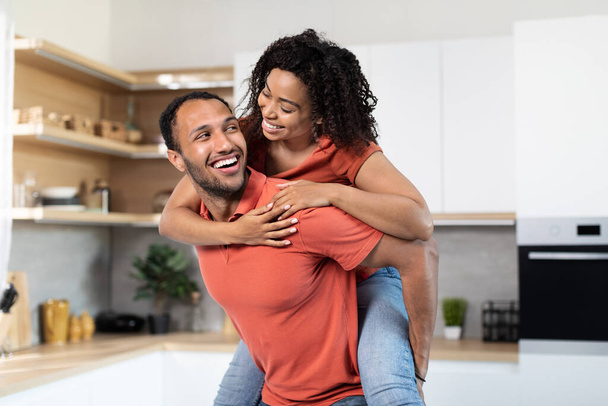 Zufriedener junger schwarzer Mann im roten T-Shirt, der die Frau auf dem Rücken hält, Spaß haben, Freizeit in der modernen Kücheneinrichtung genießen. Gemeinsames Spiel, Unterhaltung zu Hause, Paarbeziehung, Liebe und Romantik - Foto, Bild