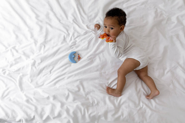 Imádnivaló afro-amerikai csecsemő baba fekszik az ágyban, és harapás fogéter, portré aranyos kis fekete fiú visel karosszéria játszik csörgőkígyó, miközben pihentető a hálószobában, felső nézet másolási hely - Fotó, kép
