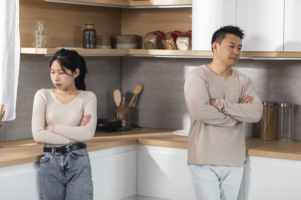 Upset korean lovers standing at home, middle eastern man and young woman with arms crossed on chest dont look at each other, having quarrel, empty space between them, kitchen interior - Photo, Image