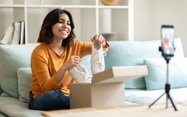 Smiling Middle Eastern Woman Recording Unboxing Video For Her Fashion Vlog, Happy Young Arab Female Unpacking Parcel With New Sneaker Shoes In Front Of Camera At Home, Using Smartphone On Tripod - Foto, imagen