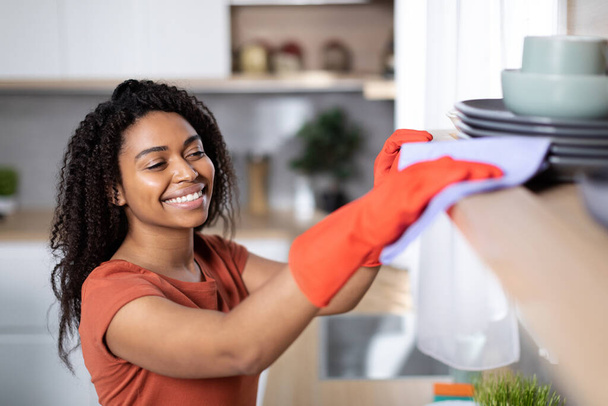 Vrolijke mooie jonge zwarte vrouw in rood t-shirt en rubberen handschoenen stof uit van houten plank in keuken interieur, kopieer ruimte. Huishoudelijke taken, perfectionisme, reinheid, hygiëne en gezondheidszorg - Foto, afbeelding