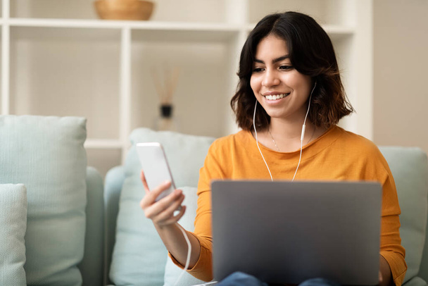 Digital Leisure. Smiling Middle Eastern Female Resting With Laptop And Smartphone At Home, Happy Young Arab Woman Wearing Earphones Sitting On Couch In Living Room And Using Modern Gaddets - 写真・画像