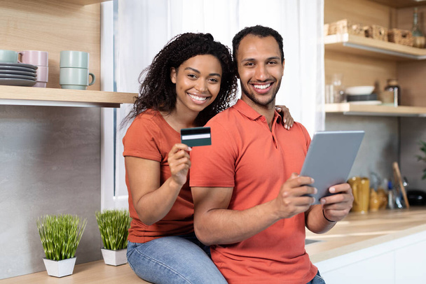 Улыбающиеся молодые чернокожие муж и жена показывают кредитную карту и планшет, наслаждаются онлайн покупками в интерьере кухни. Огромные продажи, кэшбэк, скидки, заказы товаров, любовь и отношения дома - Фото, изображение