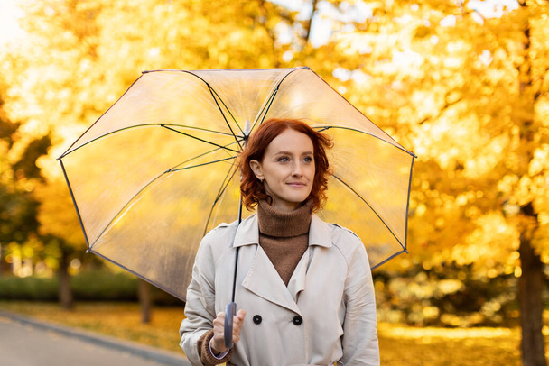 Serious pretty young european red-haired lady in raincoat with umbrella walks alone in rain in autumn in city park with yellow leaves. Free time at weekend, emotions in bad weather outdoor, covid-19 - 写真・画像