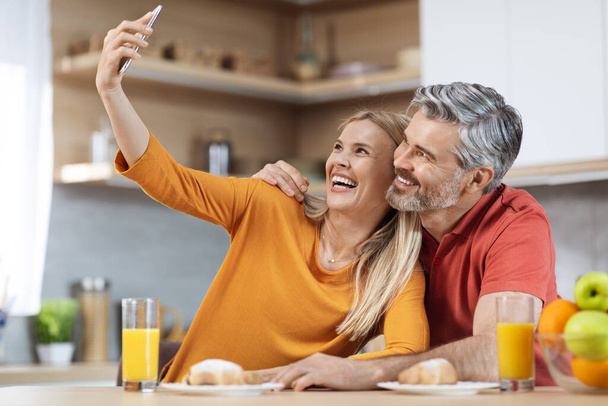 Joyeux beau couple romantique homme et femme d'âge moyen dans des vêtements confortables assis à la table de cuisine, prendre selfie tout en prenant le petit déjeuner ensemble à la maison, en utilisant un smartphone - Photo, image