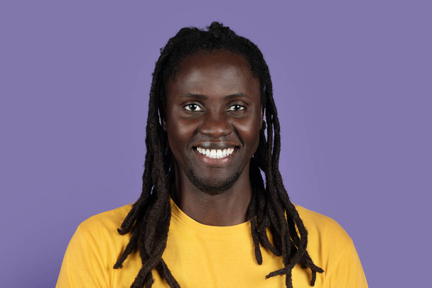 Szekrénykép egy boldog, jóképű afro-amerikai Y-generációs fickóról, hosszú copfokkal, aki lila stúdió háttér felett mosolyog, sárga pólóban. A feketék életmódjának koncepciója - Fotó, kép