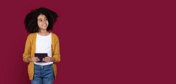 Happy preteen african american kid girl in casual with bushy hair using modern digital tablet, chatting with friends, looking at copy space over burgundy studio background, panorama - Photo, Image