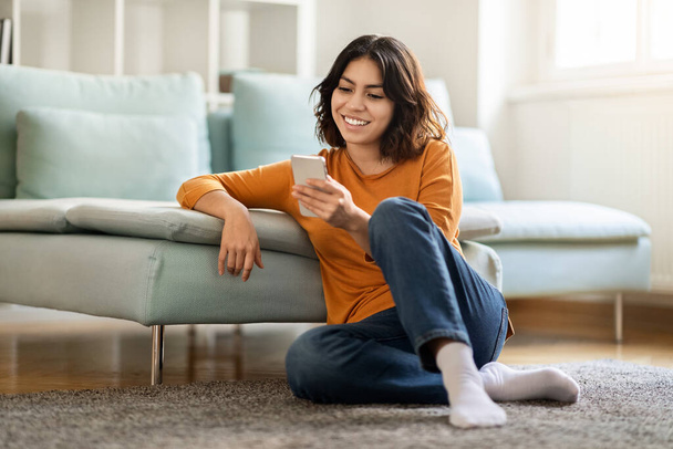 Smiling Young Arab Lady With Smartphone Relaxing On Floor At Home, Happy Beautiful Middle Eastern Female Brosing Internet On Mobile Phone Or Shopping Online While Resting In Living Room, Free Space - Foto, Imagem