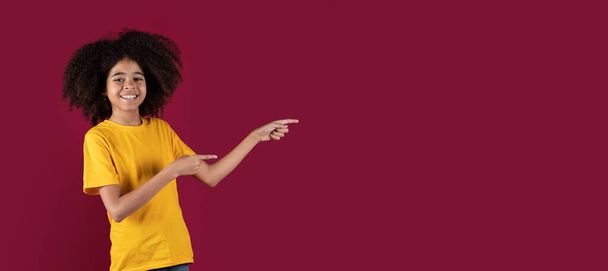 Alegre linda niña afroamericana con el pelo tupido apuntando al espacio de copia para la publicidad y sonriendo sobre fondo de estudio borgoña, mostrando algo emocionante, panorama - Foto, Imagen