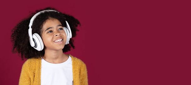 Cute african amerykańskie dziecko kręcone nastolatek dziewczyna włosy przy użyciu zupełnie nowe białe słuchawki bezprzewodowe, patrząc na przestrzeń kopiowania i uśmiechając się na tle studio bordowy, słuchając muzyki, panorama - Zdjęcie, obraz
