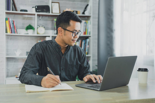 Aasialainen freelance mies yllään lasit ja hymy työskentelevät kirjoittamalla kannettavan tietokoneen ja kirjoittaa muistiinpanoja kannettavan kotona. Yrittäjä mies työskentelee liiketoiminnan olohuoneessa kotiin. Liike-elämä kotona käsite. - Valokuva, kuva