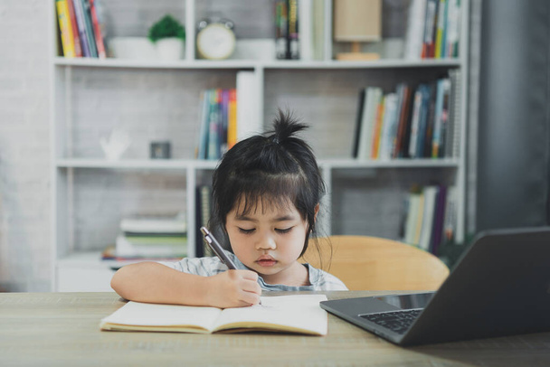 Азиатская девочка в голубой полосатой рубашке пользуется ноутбуком и пишет заметки в блокноте, чтобы учиться онлайн на деревянном столе в гостиной дома. Образование онлайн из домашней концепции. - Фото, изображение