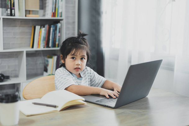 Aasialainen vauva tyttö yllään sininen raidallinen paita käyttää kannettavaa tietokonetta ja muistikirjaa opiskella verkossa puupöydällä olohuoneessa kotona. Koulutus oppiminen verkossa kotoa käsite. - Valokuva, kuva
