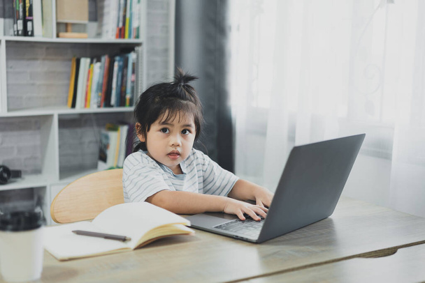 Aasialainen vauva tyttö yllään sininen raidallinen paita käyttää kannettavaa tietokonetta ja muistikirjaa opiskella verkossa puupöydällä olohuoneessa kotona. Koulutus oppiminen verkossa kotoa käsite. - Valokuva, kuva