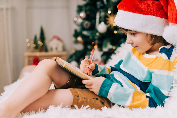 赤いサンタの帽子の子供の男の子が手紙を書いて、自宅で親愛なるサンタに絵を描く。子供の願い事リスト。クリスマスの贈り物の夢。メリークリスマスとハッピーニューイヤー - 写真・画像