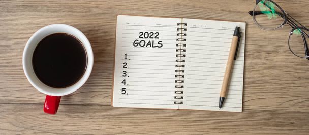 2022年目標ノートブック、ブラックコーヒーカップと木製のテーブルの上にペンで、トップビューとコピースペース。幸せな新年、解決、リスト、戦略と計画の概念を行うには - 写真・画像