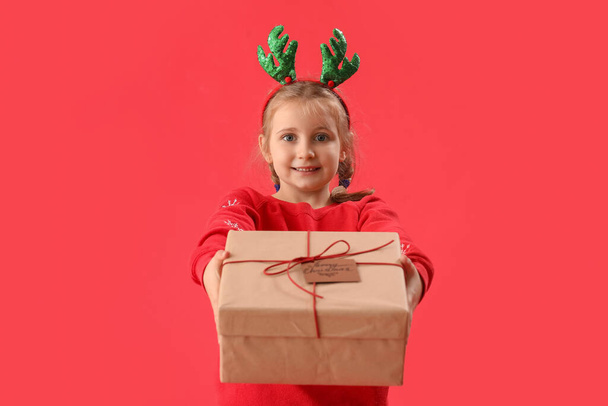Szczęśliwa dziewczynka w rogach reniferów z prezentem świątecznym na czerwonym tle - Zdjęcie, obraz