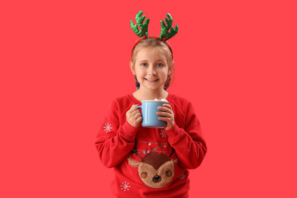Joyeux petite fille en cornes de renne avec tasse de cacao sur fond rouge - Photo, image