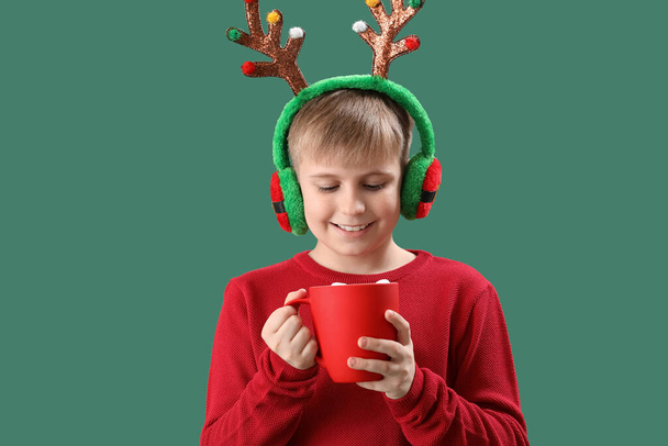 Happy little boy in reindeer horns with cup of cocoa on green background - Photo, Image