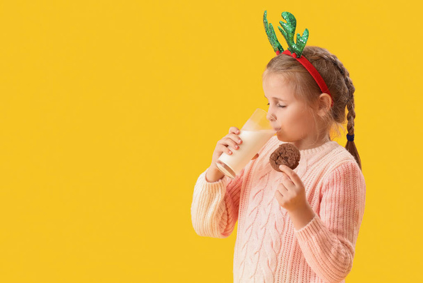 Geyik boynuzu takmış, kurabiyeli, sarı arka planda süt içen mutlu küçük kız. - Fotoğraf, Görsel