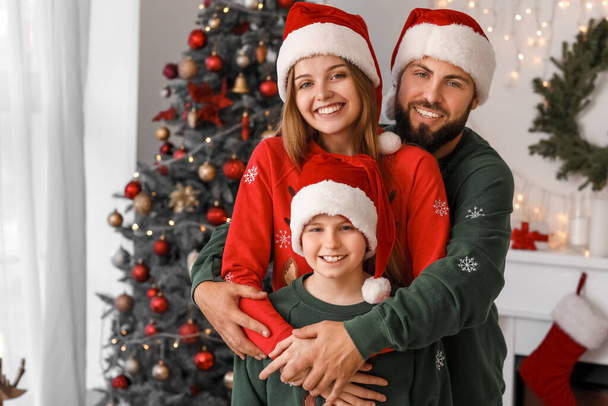 Padres felices con su hijo pequeño en los sombreros de Santa abrazándose en casa en la víspera de Navidad - Foto, Imagen