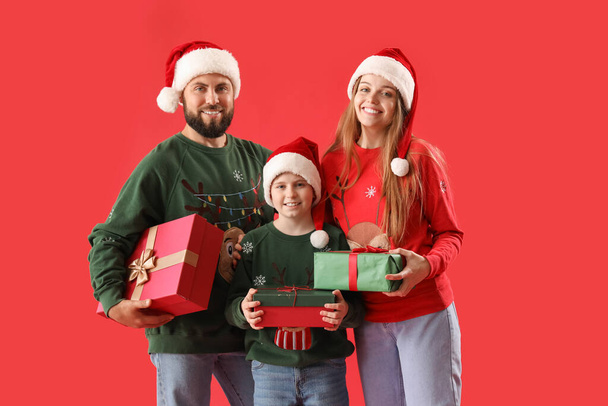 Szczęśliwi rodzice ze swoim synkiem w kapeluszach Mikołaja i prezentach świątecznych na czerwonym tle - Zdjęcie, obraz