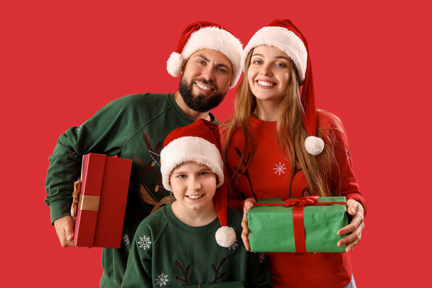 Genitori felici con il loro piccolo figlio in cappelli di Babbo Natale e regali di Natale su sfondo rosso - Foto, immagini