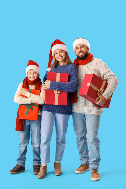 Onnelliset vanhemmat pienen poikansa kanssa Santa-hatuissa ja joululahjoissa sinisellä taustalla - Valokuva, kuva