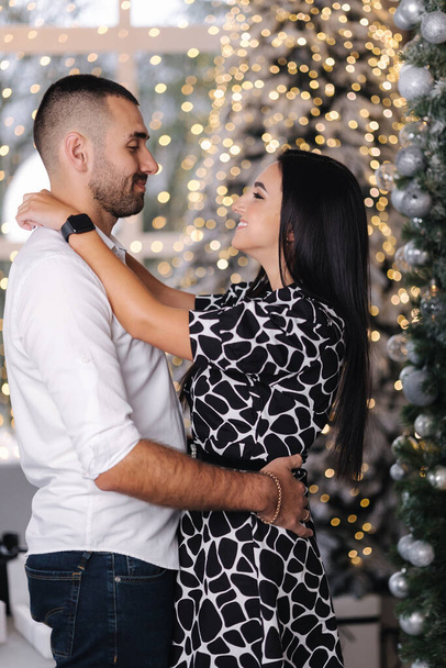 Handsome man with his beautiful woman hug each other at home at Christmas eve. Fit tree and snow outsif. Christmas mood. - Foto, Imagen