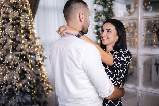 Handsome man with his beautiful woman hug each other at home at Christmas eve. Fit tree and snow outsif. Christmas mood. - Foto, immagini
