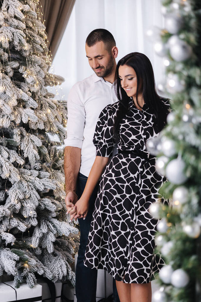 Vonzó nő gyönyörű fekete-fehér ruha állni a férje előtt fitt fa. Karácsonyi hangulat otthon. - Fotó, kép