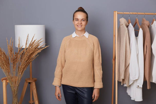 Portrait of attractive cheerful woman with bun hairstyle wearing beige sweater standing in fashion sore near clothing on hangers, looking at camera, presenting her online showroom. - Fotografie, Obrázek