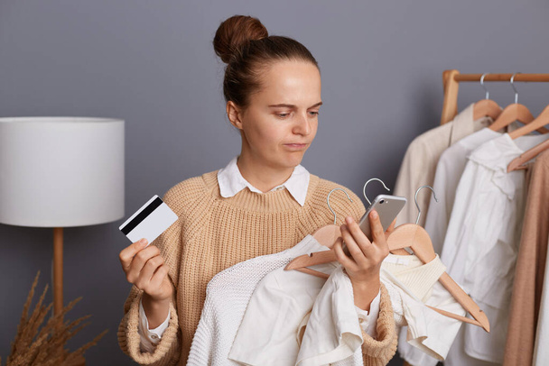 Portrait of disappointed sad Caucasian woman wearing beige jumper standing with credit card and smart phone, holding clothing on hangers, having unsuccessful payment for her new outfit. - Foto, afbeelding