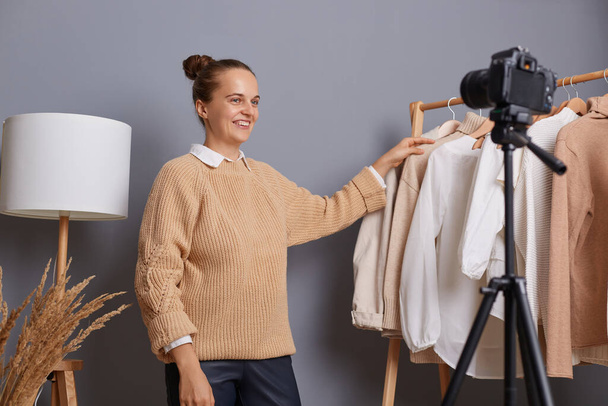 Image of good looking smiling woman fashion blogger with bun hairstyle showing basic outfits to subscribers, recording video, using camera with tripod or broadcasting livestream. - Photo, Image