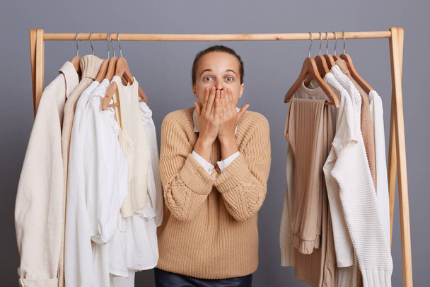 Horizontal shot of shocked Caucasian woman buyer standing near clothes hang on shelf, selling trendy outfit, being excited and surprised, covering mouth with palms. - Photo, Image