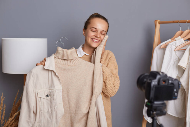 Image of smiling pretty pleased female with bun hairstyle recording video for her followers, using camera with tripod, showing soft sweater on hangers. - Foto, immagini