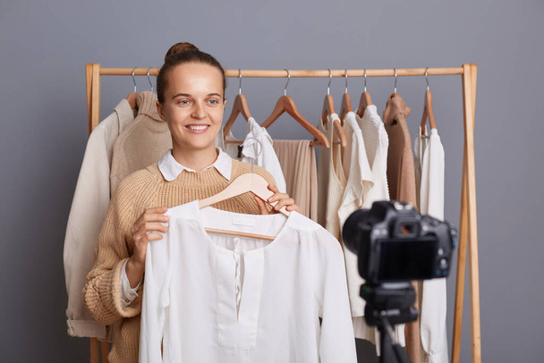 Indoor shot of pretty pleased positive woman with bun hairstyle recording video on camera, holding white shirt on hanger, showing fashionable look for her female followers. - Foto, afbeelding