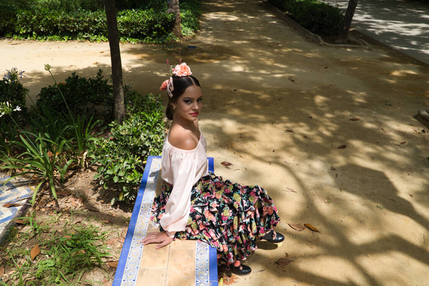 Красивая танцовщица фламенко в цветущей юбке с оборками и цветами в волосах сидит на скамейке в парке, улыбаясь и позируя для фотографий.. - Фото, изображение