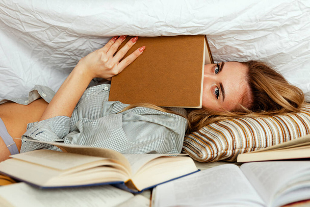 Roztomilá blondýnka v bílém tričku na posteli v domácí ložnici, čte knížku. Model pod přikrývkou s knihou. Mnoho knih v popředí - Fotografie, Obrázek