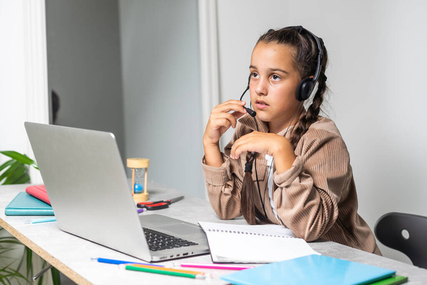 Χαμογελώντας λίγο Καυκάσιος κορίτσι στα ακουστικά handwrite μελέτη σε απευθείας σύνδεση χρησιμοποιώντας το φορητό υπολογιστή στο σπίτι, χαριτωμένο ευτυχισμένη μικρό παιδί στα ακουστικά να λάβει το Διαδίκτυο web μάθημα ή τάξη στον υπολογιστή, κατ 'οίκον διδασκαλία έννοια. - Φωτογραφία, εικόνα