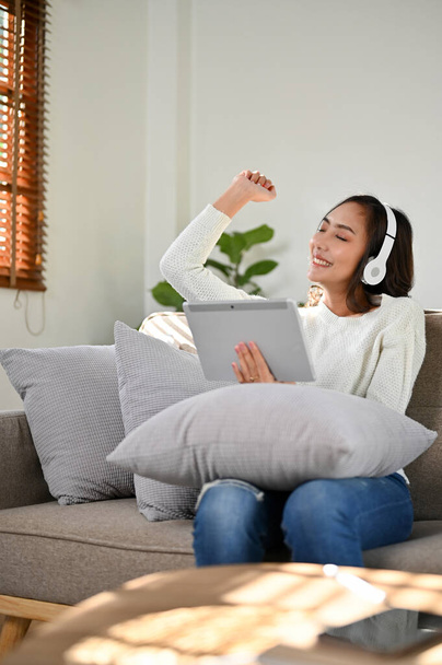 Portret, Relaksująca i radosna milenijna Azjatka w luźnych ciuchach nosząca słuchawki, słuchająca muzyki, korzystająca z tabletu, siedząca na kanapie w salonie. koncepcja czasu wolnego i stylu życia - Zdjęcie, obraz