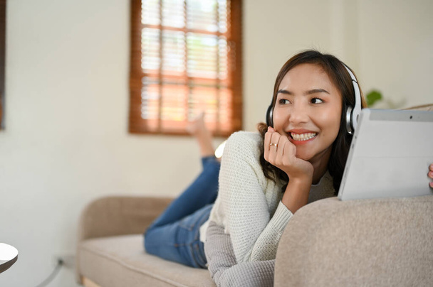 Mulher asiática jovem atraente em roupas casuais usando fones de ouvido, usando tablet, sorrindo e olhando para longe da câmera enquanto deitado no sofá na sala de estar. - Foto, Imagem