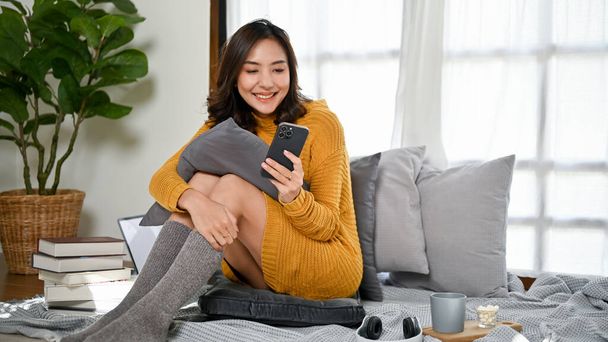 Attrayant froid jeune femme asiatique en pull confortable et chaussettes assis dans son salon et en utilisant son smartphone. SMS, lecture de blog en ligne, vérification des médias sociaux, mise à jour de l'application mobile - Photo, image