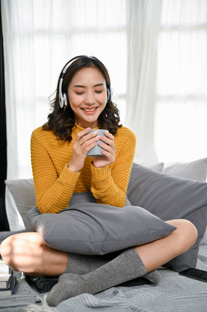 Relaxed and happy young Asian woman in comfy sweater and socks wearing headphones, listening to music and enjoying morning coffee on her comfortable bed in her bedroom. - Photo, image