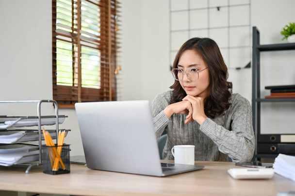 Inteligentna i profesjonalna milenijna azjatycka bizneswoman lub menedżerka koncentrująca się na swoich zadaniach, patrząca na ekran laptopa, myśląca i planująca nową strategię dla biznesu, pracująca w biurze. - Zdjęcie, obraz