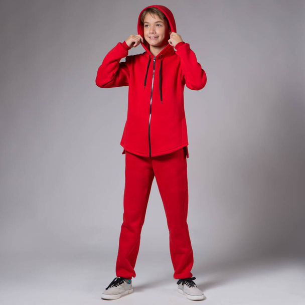Un adolescente en un chándal rojo, sonriendo, se para sobre un fondo gris, endereza su capucha con ambas manos - Foto, Imagen