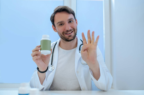 Waist-up portrait of a joyful general practitioner with the plastic vitamin bottle in his hand during the consultation - Foto, Bild